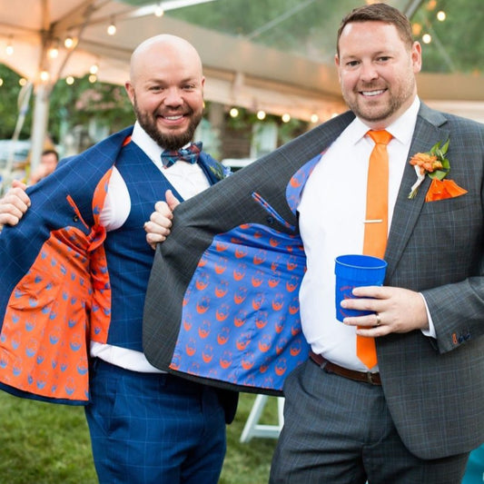 Two men in suits holding up a colorful jacket with patterns, standing outdoors under a tent.
